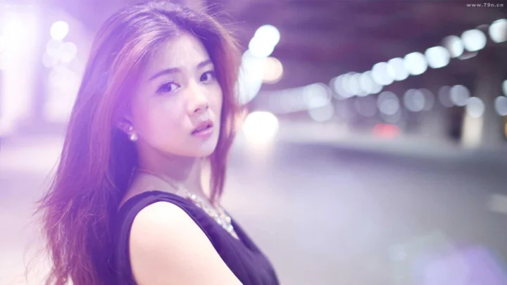 一位长发女性身穿黑色无袖上衣，站在紫色光影与模糊城市灯光前。
