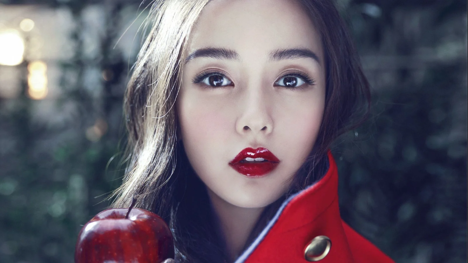 一名成年女子穿着红色外套，在暗色树木背景前手持红苹果。