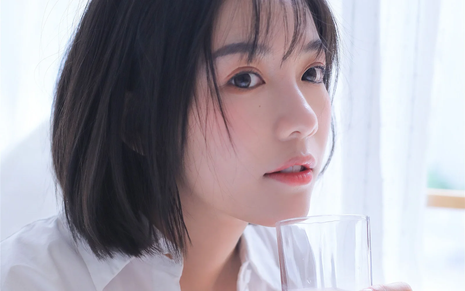 一位短发女子在明亮窗边手持玻璃杯凝望镜头。