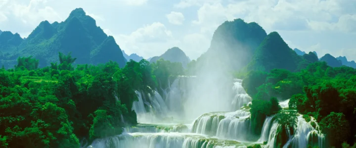 青翠山林之间，层叠瀑布从群山间流淌而下，薄雾弥漫在明亮的天空下。