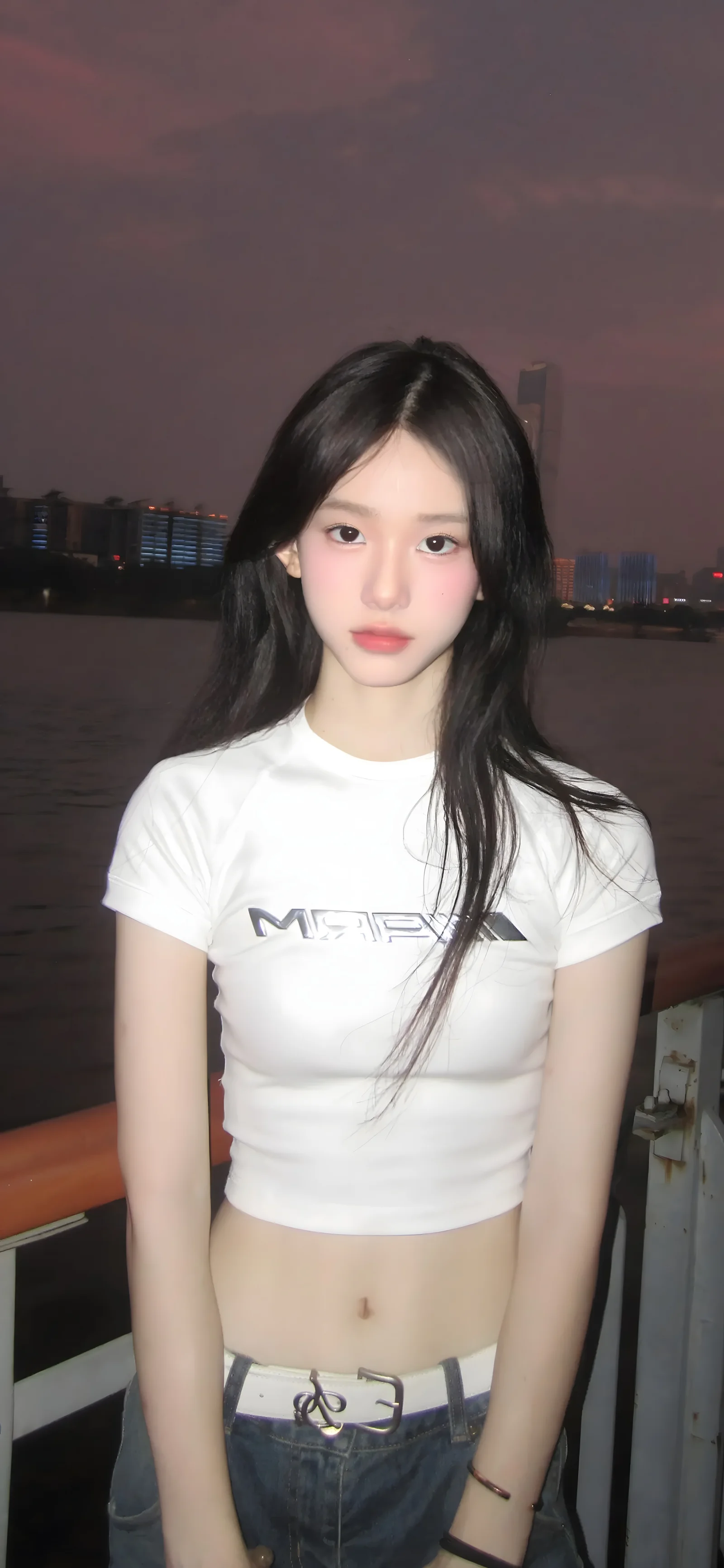 一名长发女子穿着白色上衣站在夜色中的江畔栏杆旁，远处可见城市灯光。