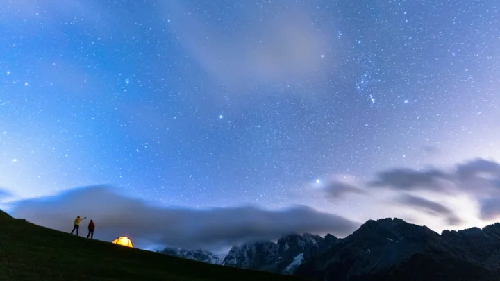 繁星点点的夜空笼罩着云雾缭绕的深色山脉，山坡上可见一顶亮着灯的帐篷。