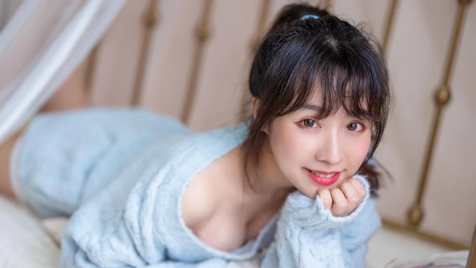 검은 머리의 여성이 연한 파란색 스웨터를 입고 침대에 엎드려 카메라를 부드럽게 바라보고 있다.