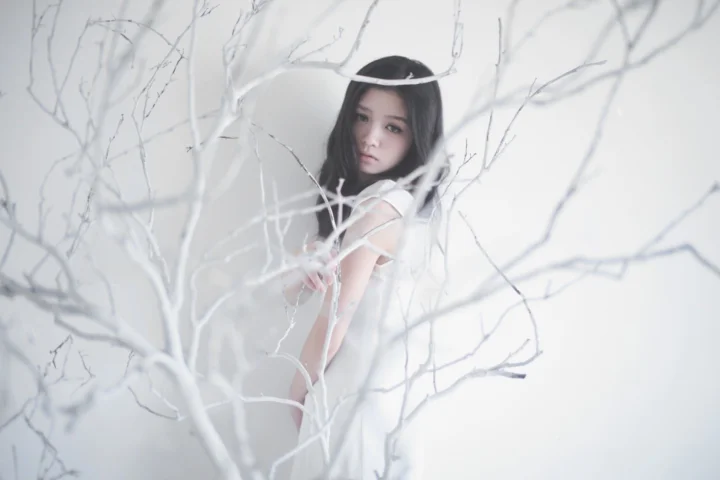 一名身穿白色服装的成年女子站在白色枝条之间，画面以浅色调为主。