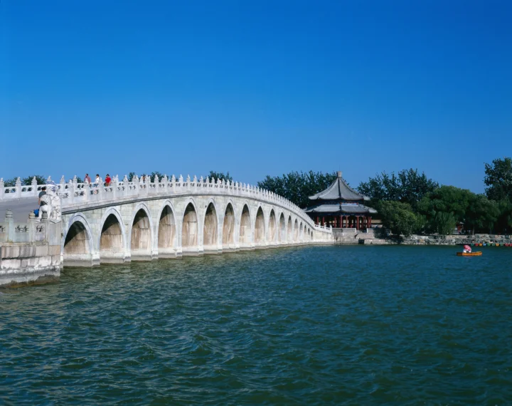 蓝天下，一座白色拱桥横跨湖面，远处可见传统亭台与绿树。