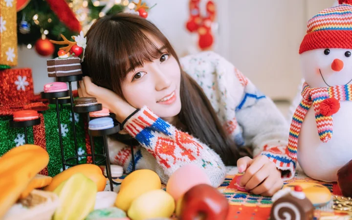 女性がクリスマスツリーや飾り、雪だるまのそばでテーブルに寄りかかりながら微笑んでいる。