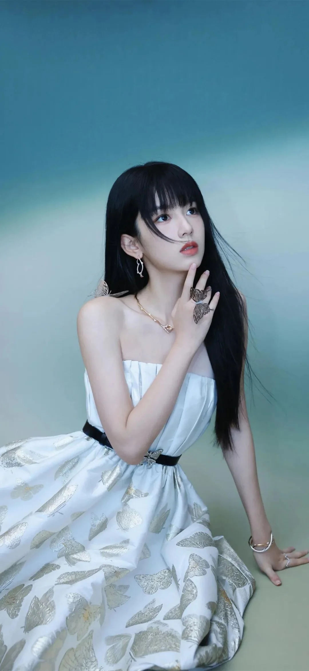 一位长发女性身穿白色图案连衣裙，坐在蓝色渐变背景前，手指轻触脸颊。