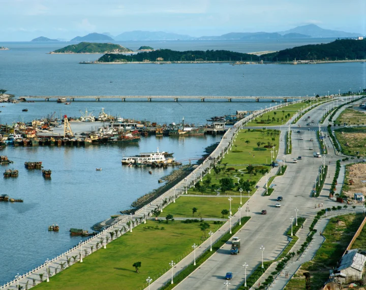 画面展示了海港、停泊的船只、沿海道路、绿地以及远处的岛屿和山峦。