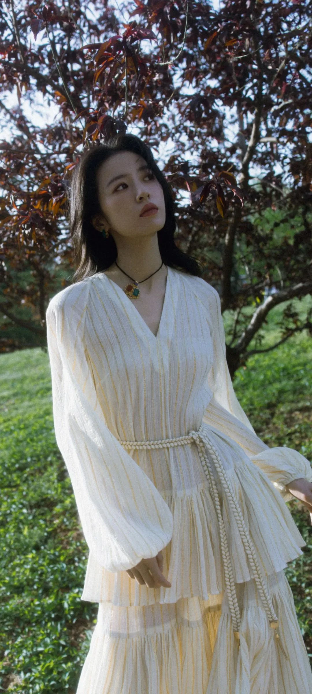 흰색 긴소매 원피스를 입은 성인 여성이 나무로 둘러싸인 야외에 서 있다.