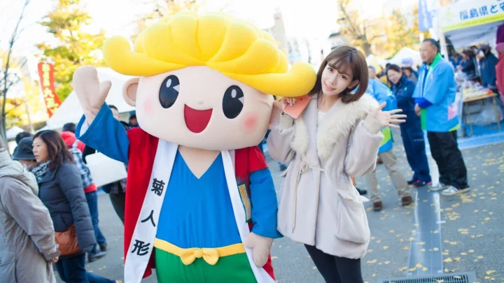 파란색과 빨간색 의상을 입은 마스코트와 여성이 야외 거리 행사 인파 속에 서 있다.