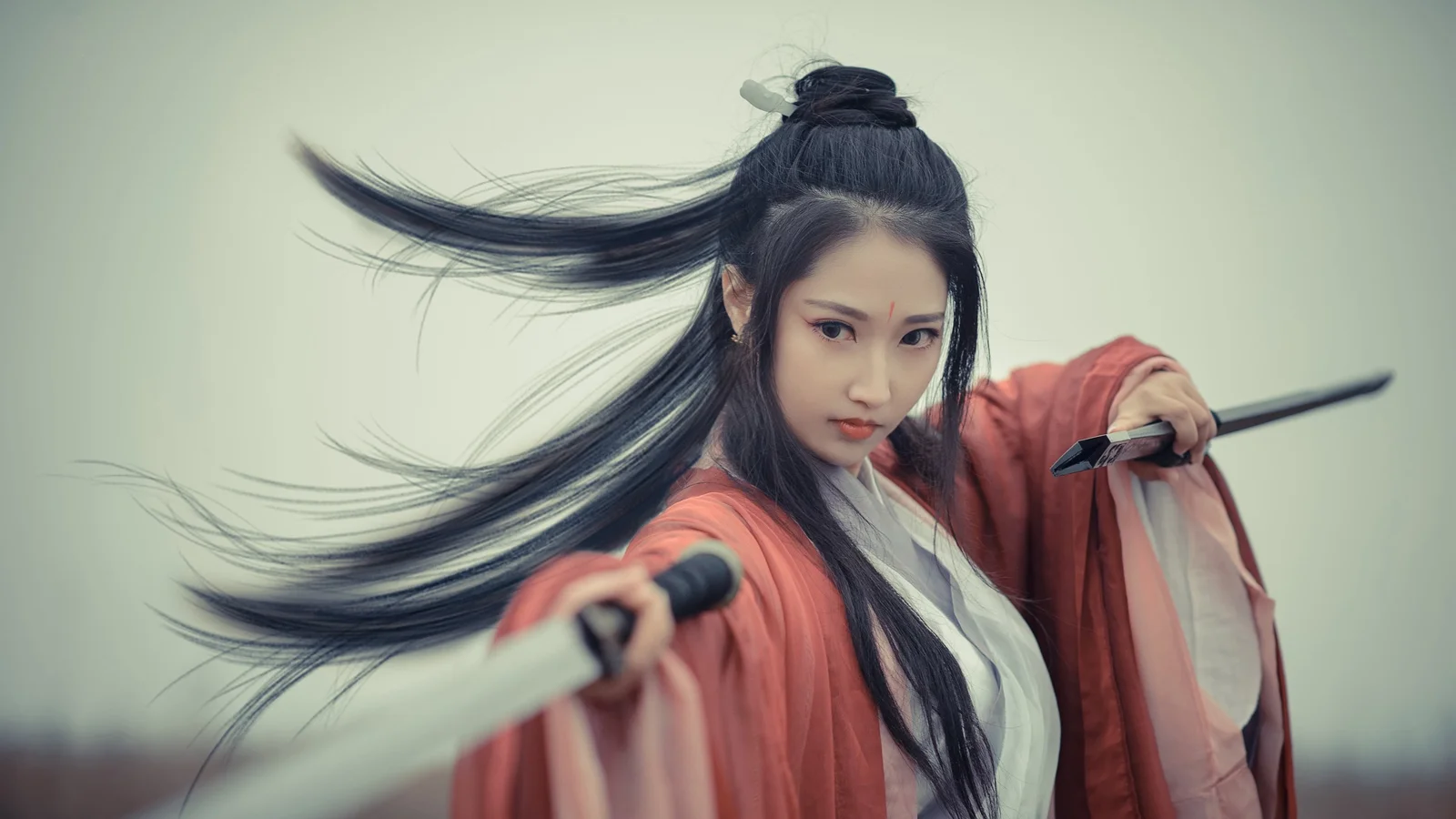 붉은 전통 의상을 입은 긴 머리 여성이 두 자루의 검을 들고 카메라를 바라보고 있다.