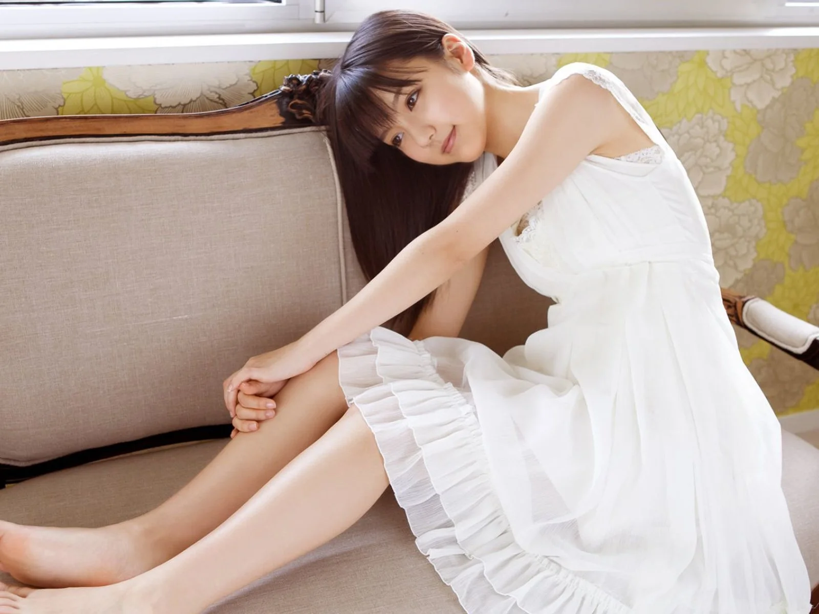 一名身穿白色连衣裙的女子坐在沙发上，室内窗边光线明亮。