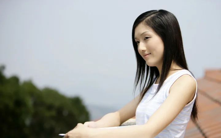 一名身穿白色无袖上衣的女子坐在栏边，身后是虚化的树木和屋顶。