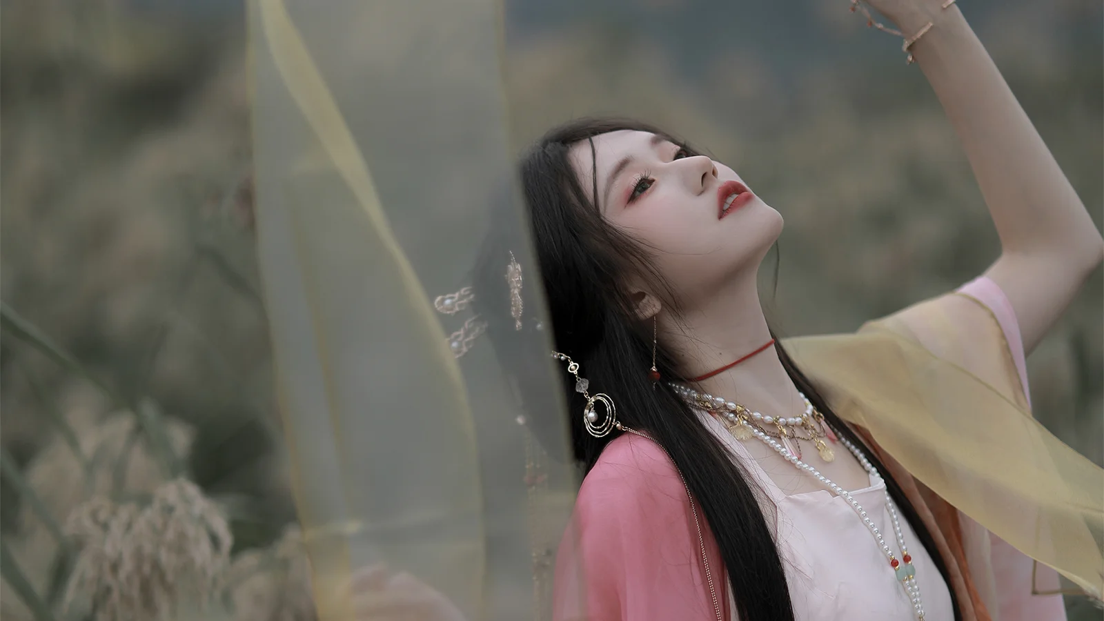 분홍색과 노란색 전통 의상을 입은 성인 여성이 야외에서 반투명한 천에 둘러싸여 고개를 들고 포즈를 취하고 있다.