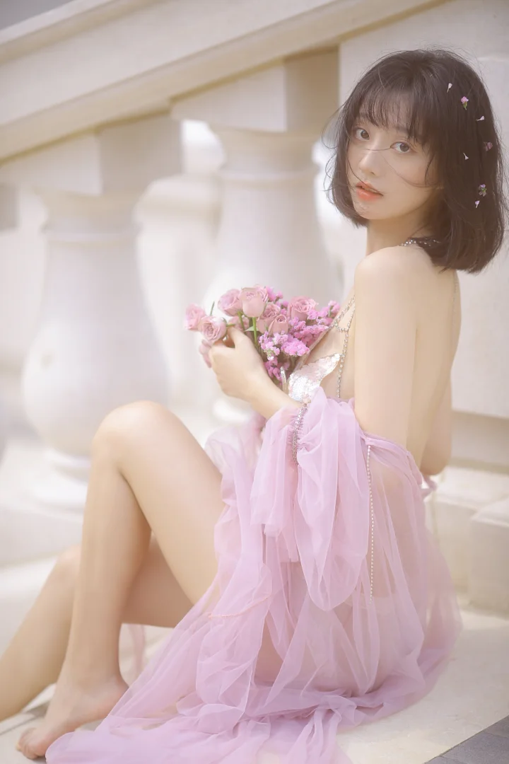 一名身穿粉色薄纱礼服的女性坐在浅色建筑柱廊旁，手捧一束粉色花朵。