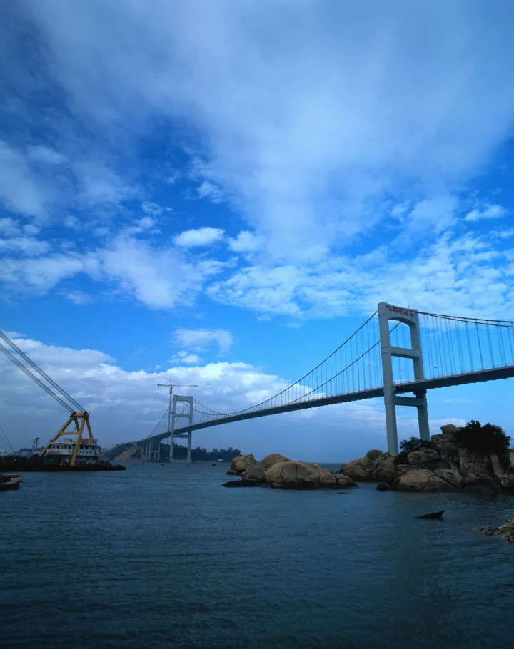 一座悬索桥横跨海面，蓝天、云层和岸边岩石构成画面主体。