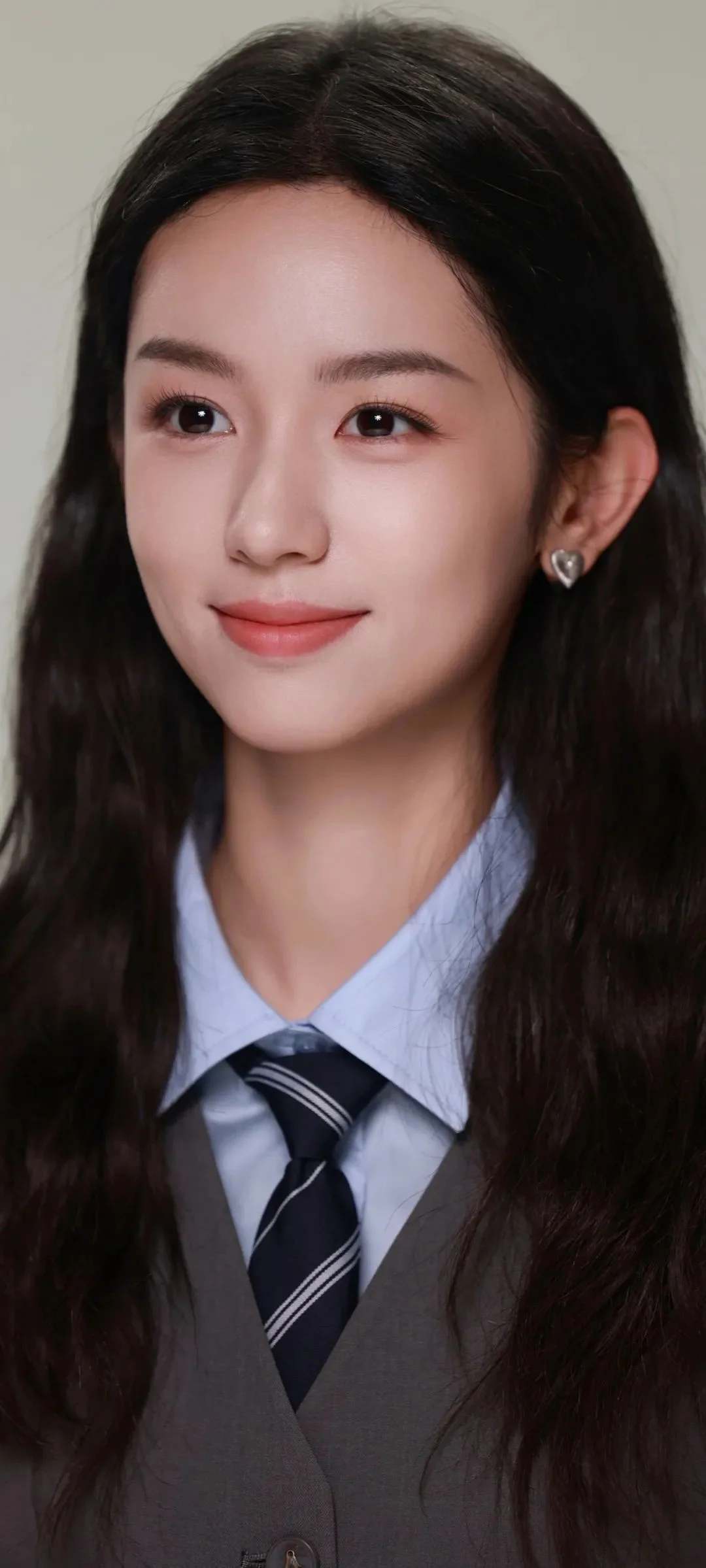 一名长发女性身穿浅蓝色领衬衫、深色领带和灰色背心，对着镜头微笑。