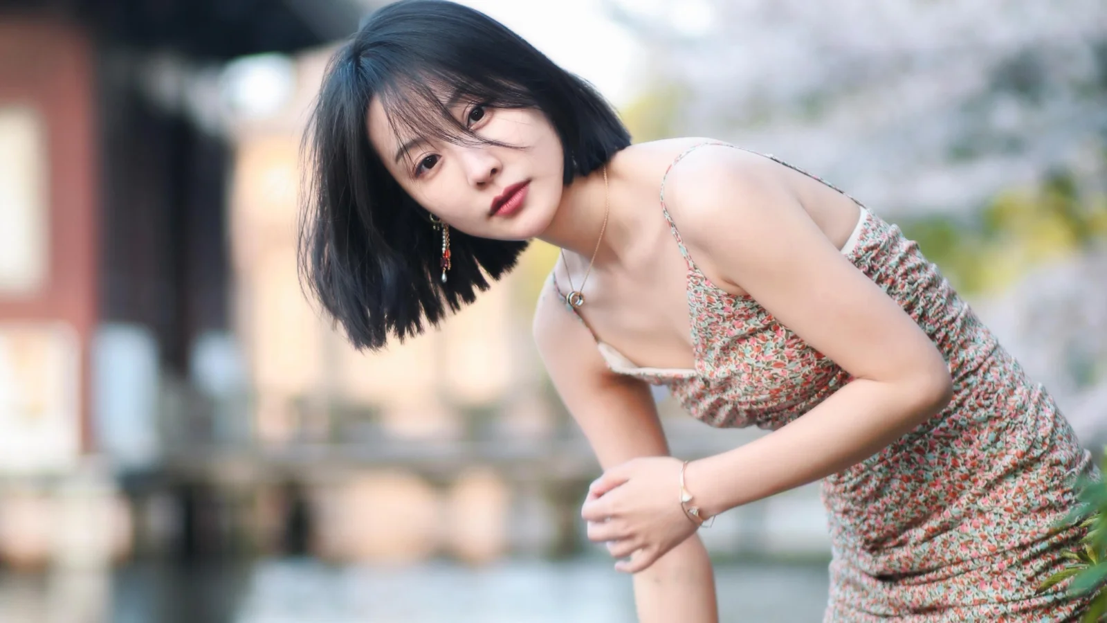 花柄のワンピースを着たショートヘアの女性が、水辺で身をかがめてカメラを見つめています。