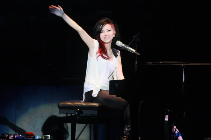 一位女性坐在钢琴前对着麦克风演奏，舞台背景昏暗。