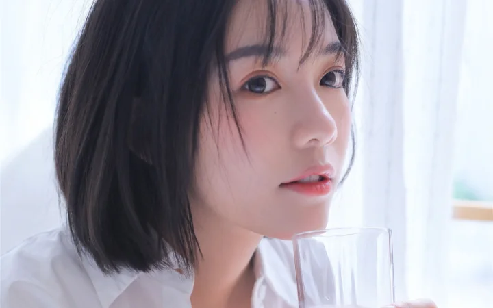 一位短发女子在明亮窗边手持玻璃杯凝望镜头。