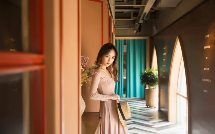 一位身穿浅色长裙的成年女性站在带有拱形窗和蓝绿色窗帘的室内走廊中。