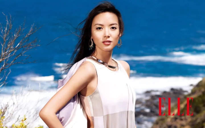 一名身穿白色连衣裙的成年女子站在蓝色海岸前，远处可见海浪与岩石。
