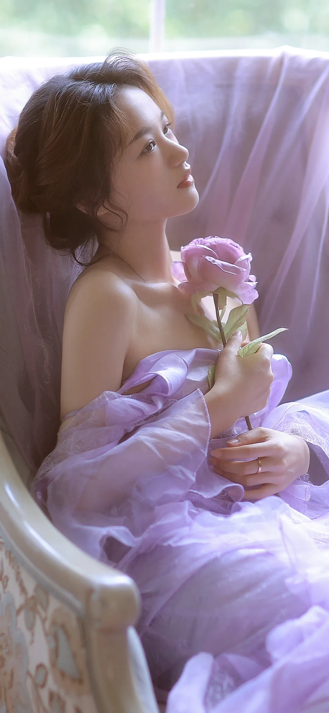 一名成年女子裹着淡紫色薄纱，手持一朵玫瑰坐在柔光窗边。