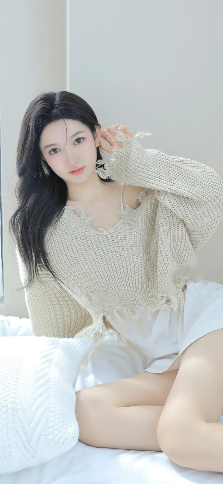 一名成年女性穿着米色宽松针织衫和白色裙子坐在床上，面向镜头摆姿势。