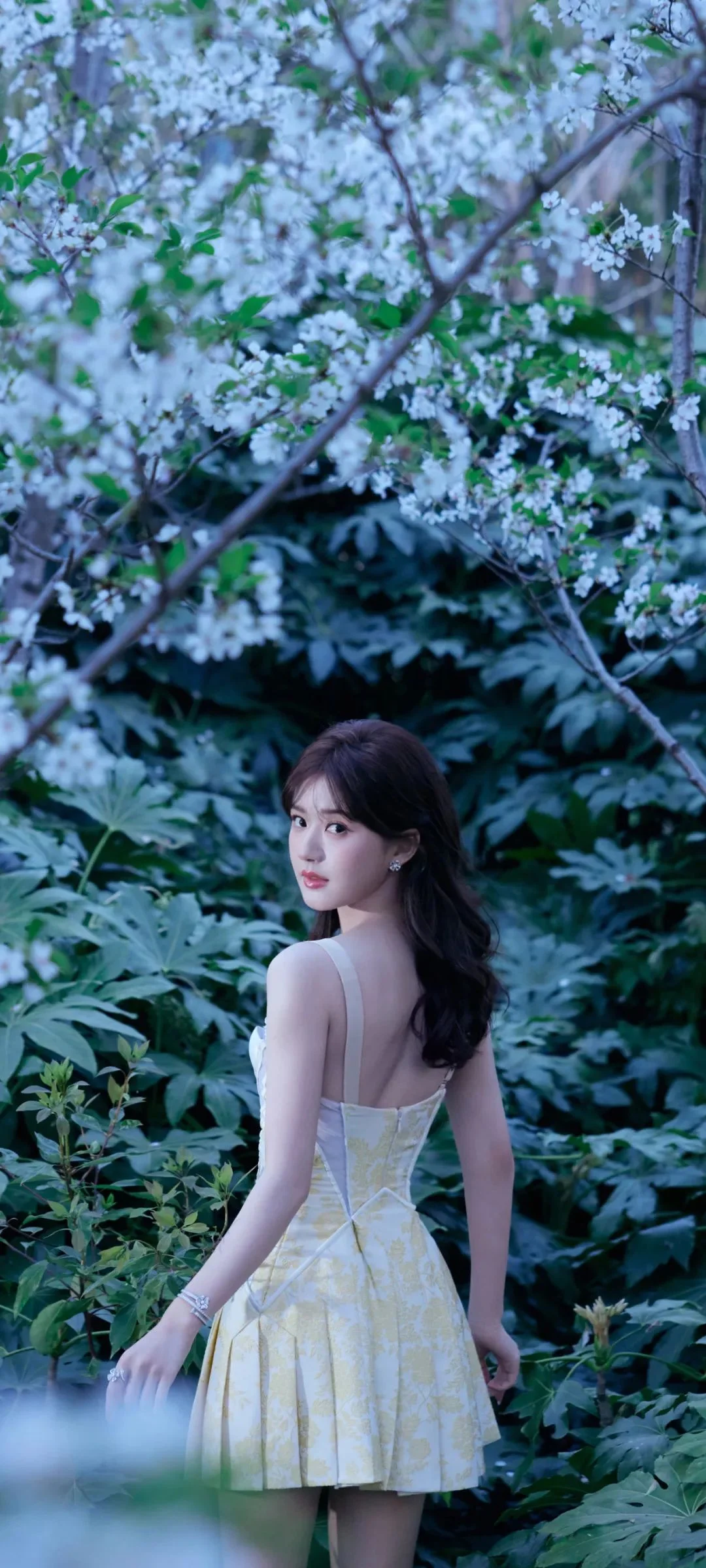 一名成年女性站在繁茂的绿叶和白色花朵之间回望镜头。