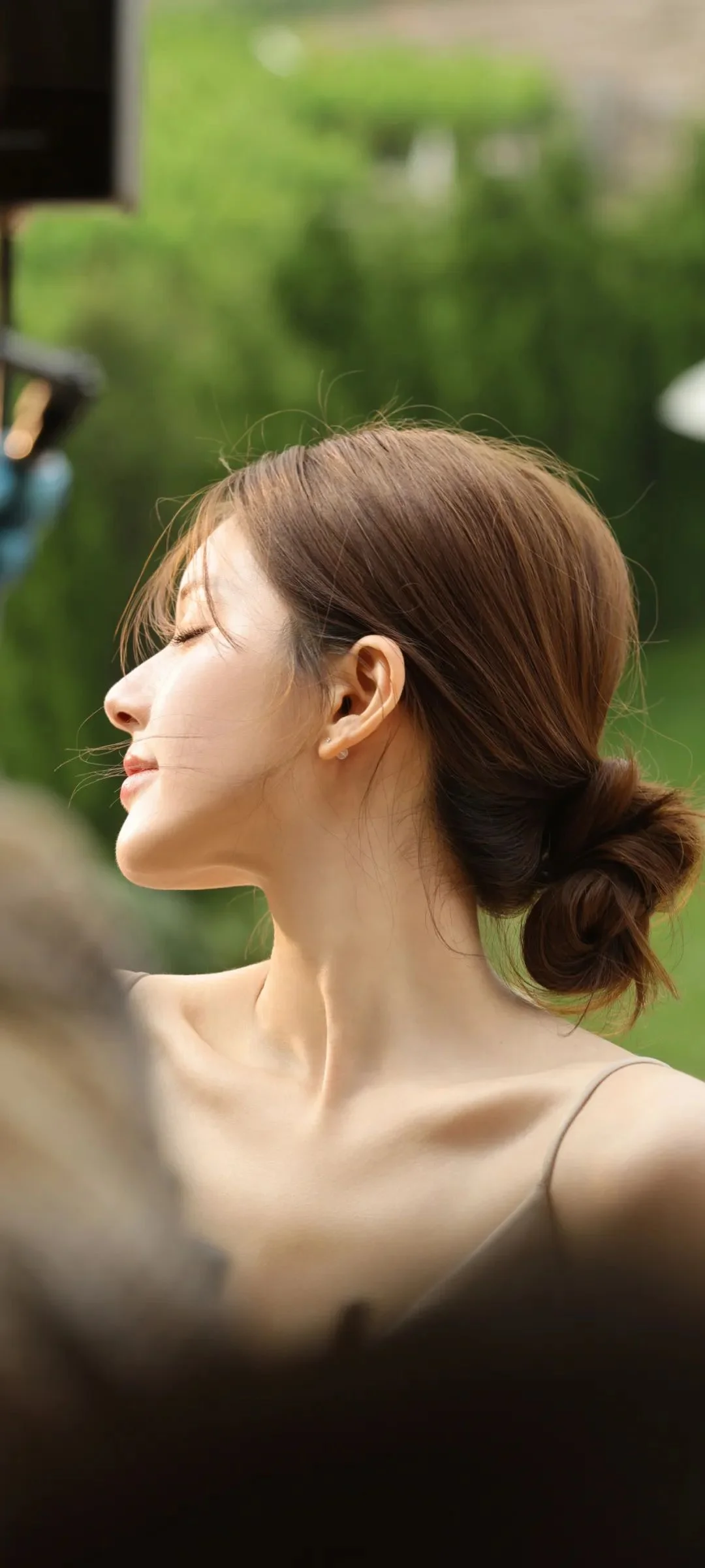 緑のぼけた屋外背景の前で、陽光に照らされた髪をまとめた成人女性の横顔を捉えています。