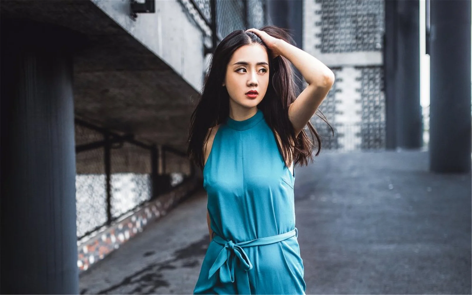一名穿蓝色无袖连衣裙的成年女子站在城市建筑之间，抬手整理头发。