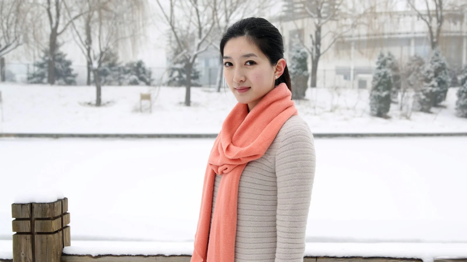 一名成年女子站在积雪覆盖的户外环境中，佩戴珊瑚色围巾。