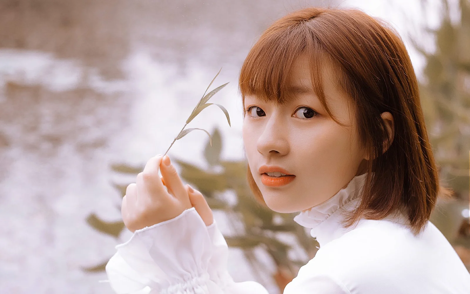 一名棕色短发女子穿着白色上衣，手持细枝站在水畔。