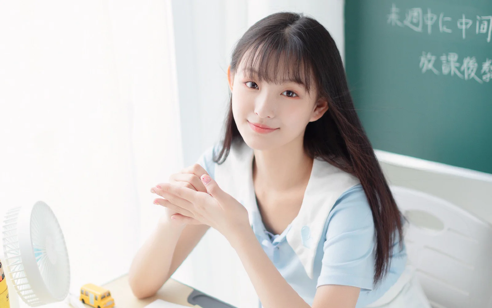 一位长发女子坐在教室里，面带微笑并将双手交叠放在身前。