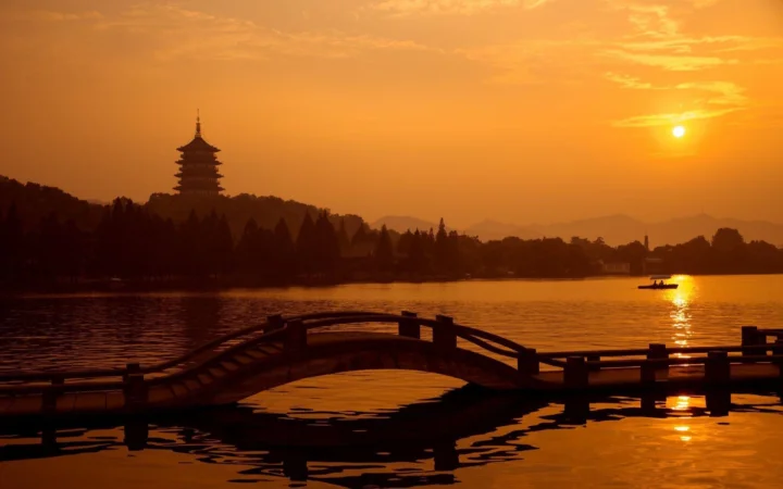 夕阳映照湖面，远处的古塔、树林与拱桥构成宁静的暮色景观。
