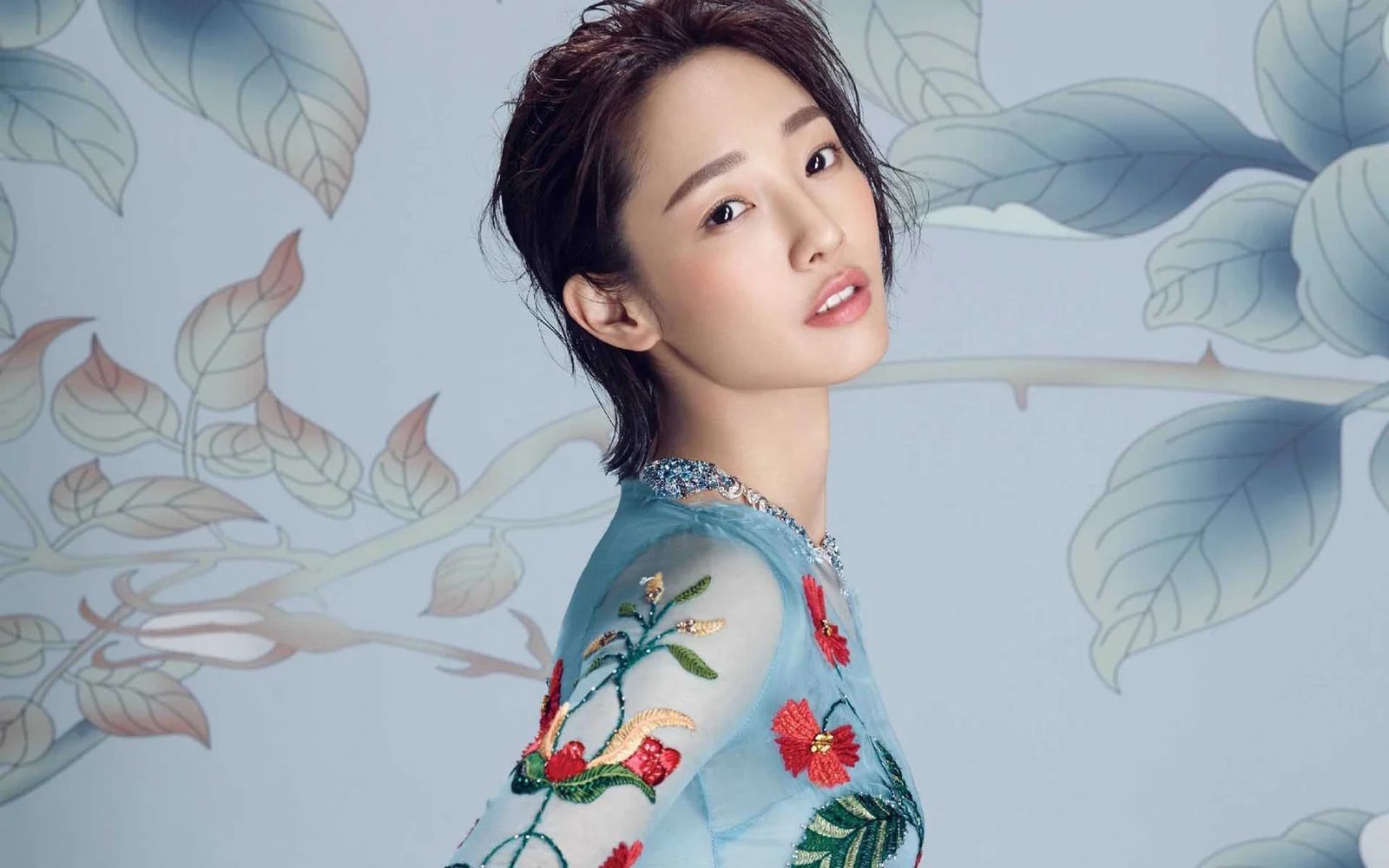 파란색 자수 의상을 입은 성인 여성이 꽃무늬 배경 앞에서 카메라를 바라보고 있습니다.