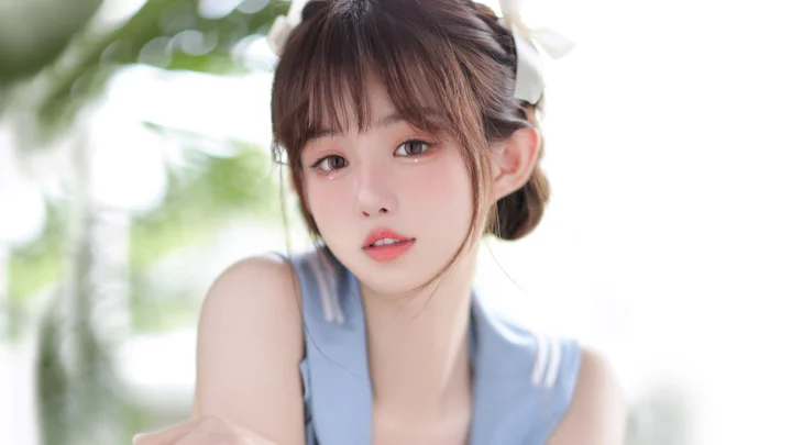 一名身穿浅蓝色上衣的女性在柔和自然光下靠近镜头，背景为虚化的绿色植物。