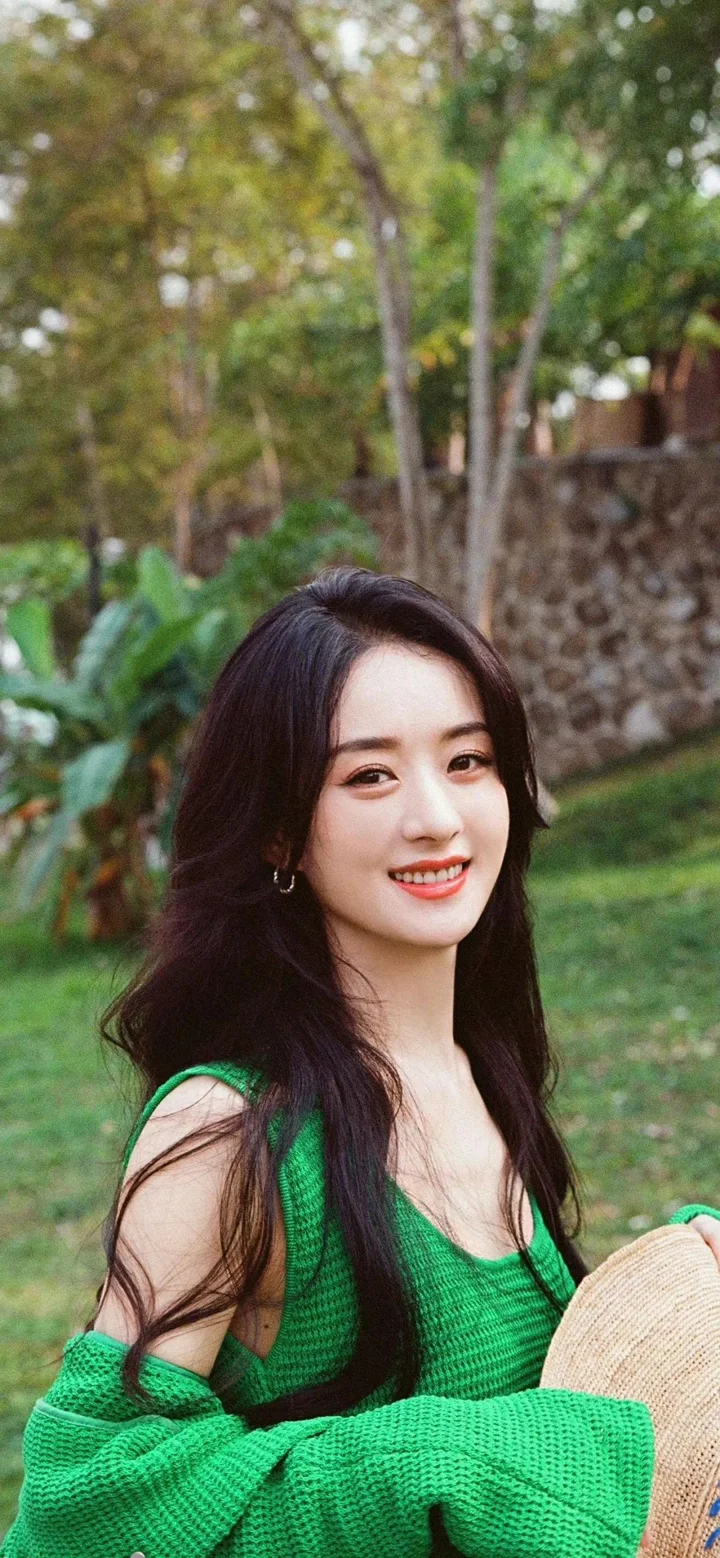 一名长发女子身穿绿色针织上衣，在户外花园中微笑。