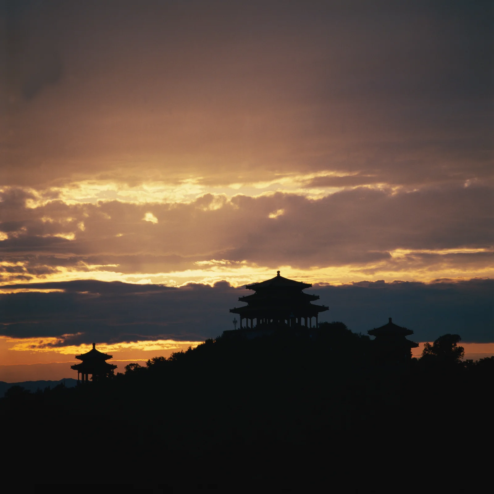 夕阳穿过层叠云层，映照出山丘上古亭建筑的深色剪影。