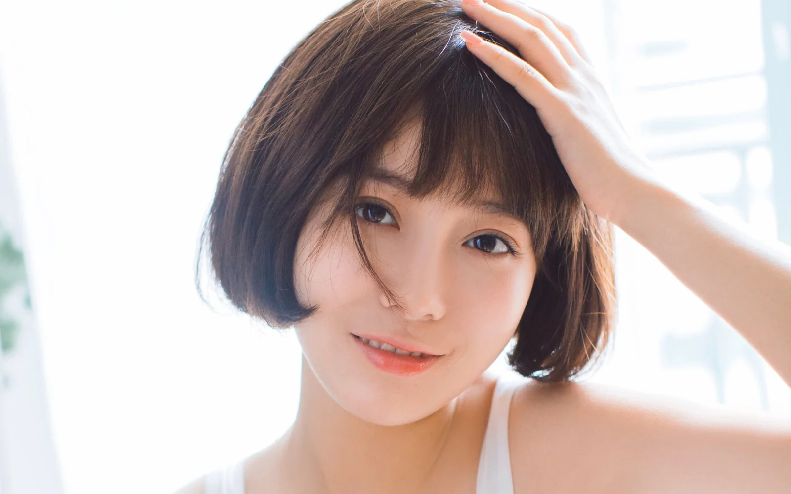 一位短发女性在明亮的窗边直视镜头，手轻触头发。