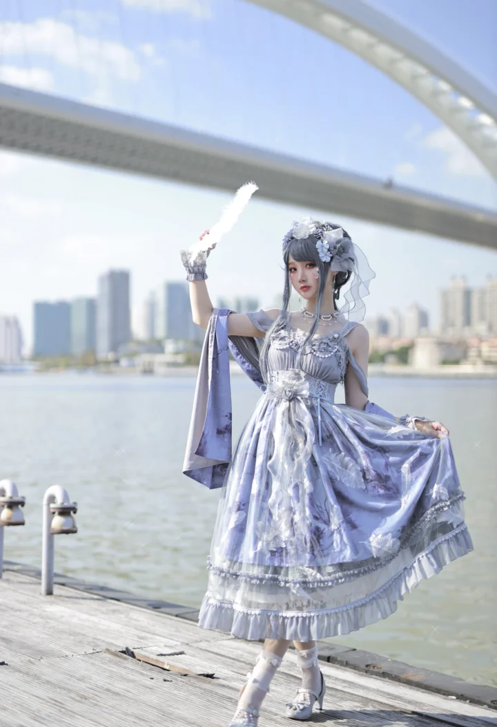 一名身穿银紫色华丽服装的女性站在水畔步道上，身后是桥梁和城市天际线。