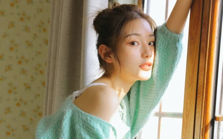 一名成年女性站在室内窗边，身穿浅绿色针织衫，阳光从窗外照入。