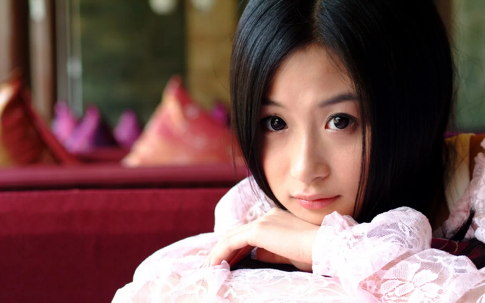 一名黑发女子身穿粉色蕾丝上衣，双臂交叠靠在沙发上面对镜头。