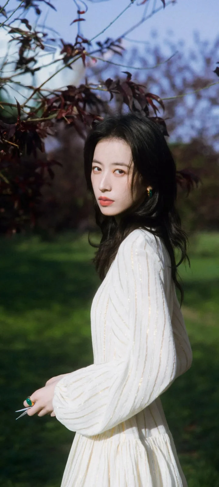 一名黑发女子穿着白色长袖连衣裙站在树木和草地之间。