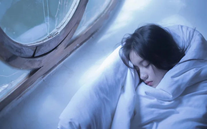 一名女子裹着白色被褥睡在窗边，画面笼罩在冷蓝色光线中。