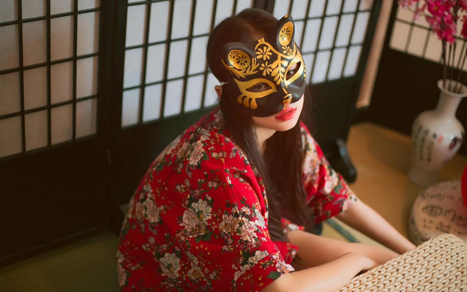 一名戴金色面具、穿红色花纹和服的女子坐在室内窗边。