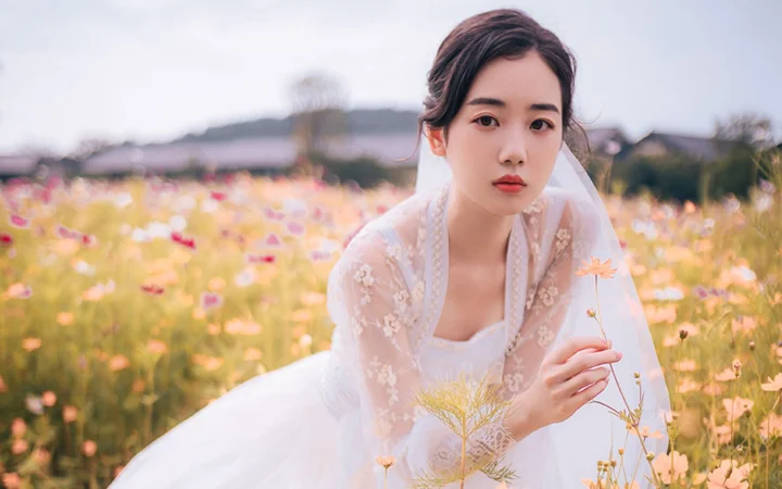 흰색 드레스를 입은 성인 여성이 꽃밭에 서서 작은 꽃을 들고 있습니다.