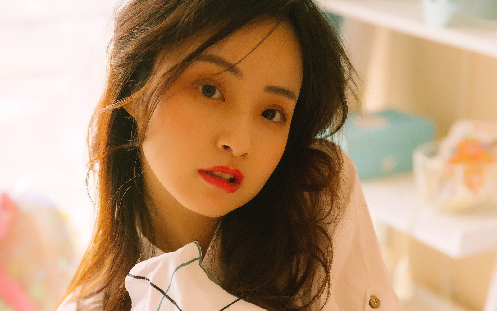 긴 검은 머리의 여성이 따뜻한 실내 조명 아래에서 빨간 립스틱을 바르고 카메라를 바라보고 있습니다.