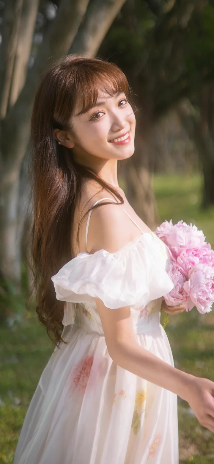 一名成年女子身穿白色连衣裙，在树木背景前手捧粉色花束回眸微笑。