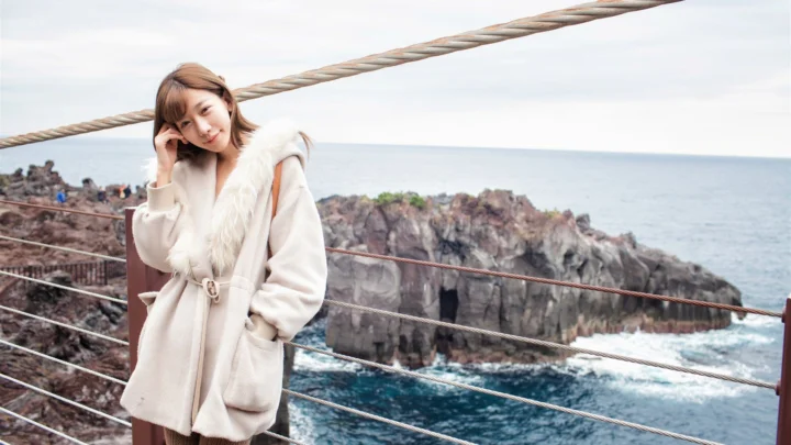 一名穿着浅色外套的女子站在海岸观景台上，身后是岩石海崖与海洋。